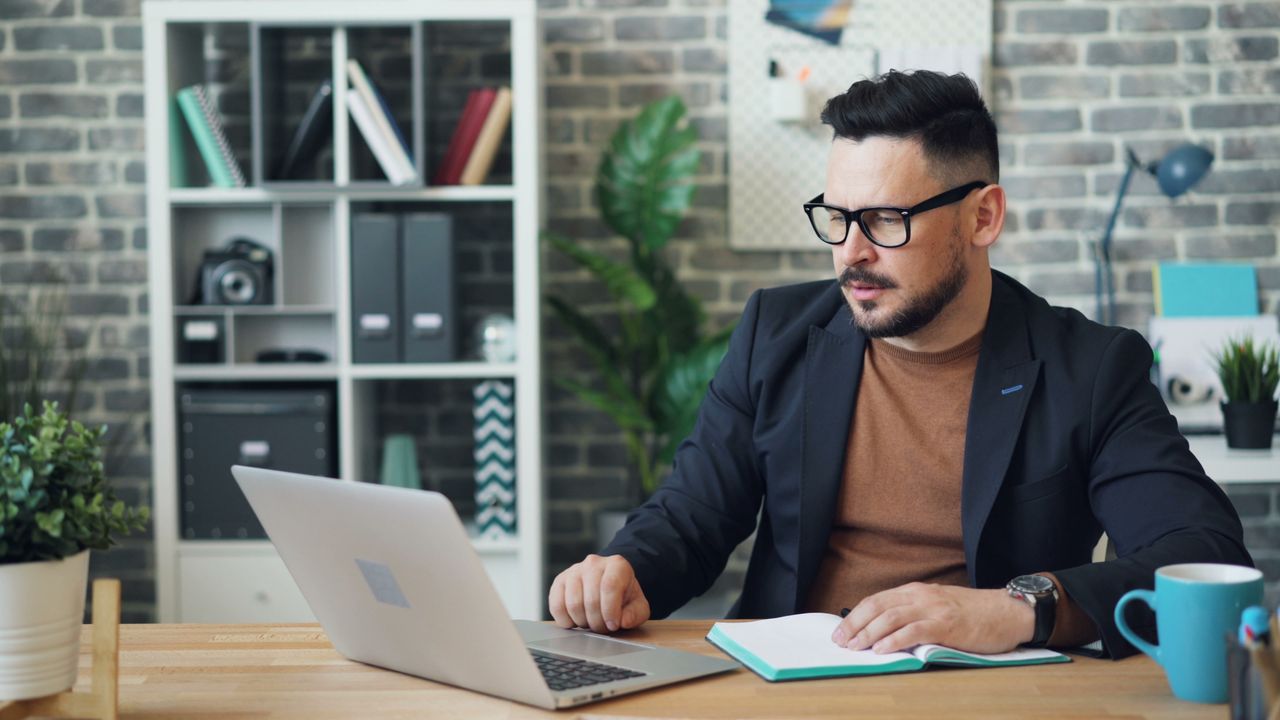 A man in a modern office wearing glasses works on a laptop surrounded by plants and bookshelves.