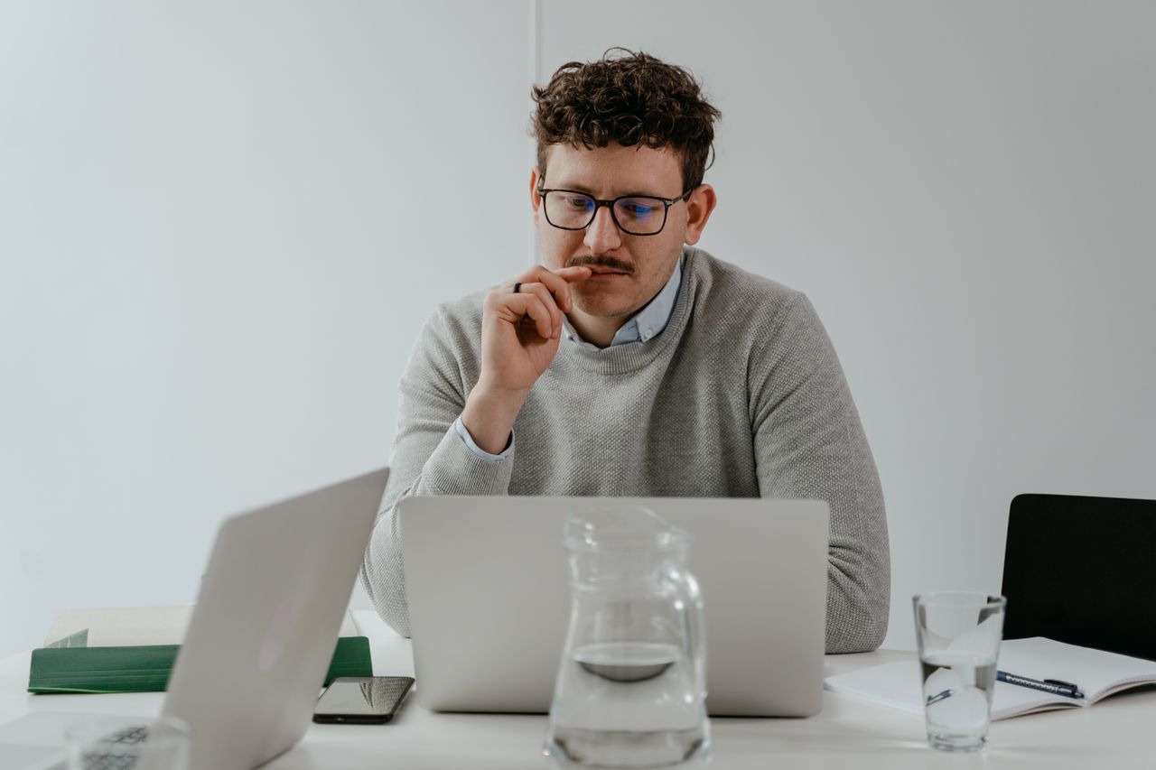 Man in glasses attentively working on a laptop at a workspace with papers and water on the table.